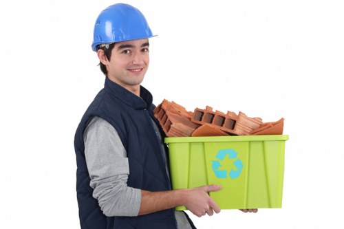 A young man wearing a blue safety helmet, navy blue vest, and grey long-sleeved shirt, standing against a plain white background and holding a bright green recycling bin with a blue recycling symbol on the front. Inside the bin are several red bricks, stacked and leaning slightly to one side, made of terracotta material with a rough texture and rectangular shape. The bricks have visible holes and ridges consistent with construction debris. The man is smiling and looking directly at the camera, presenting the bin clearly. The overall scene suggests a professional waste removal service specializing in construction waste collection, relevant to areas such as Ruislip, with a focus on recycling and rubbish disposal. The environment appears to be a studio setting, isolated from any specific outdoor or residential context, emphasizing the subject and the waste materials.
