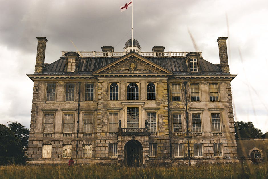 A large, historic stone building with a symmetrical façade, featuring multiple rectangular windows with cream-colored frames and decorative stone lintels. The structure displays a neoclassical archit