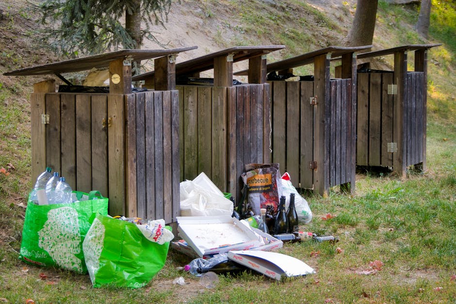 Four wooden outdoor waste bins with slanted roofs are lined up on a grassy area, positioned against a sloped background with trees and foliage. Each bin is constructed from vertical wooden planks in n